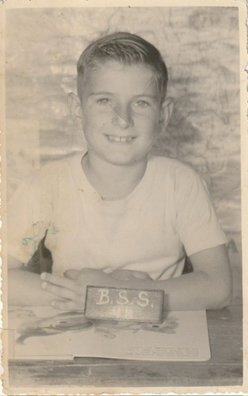 Greg Kidd in Primary School sits for a photo at a desk in a white T shirt, Brighton State School circa 1958